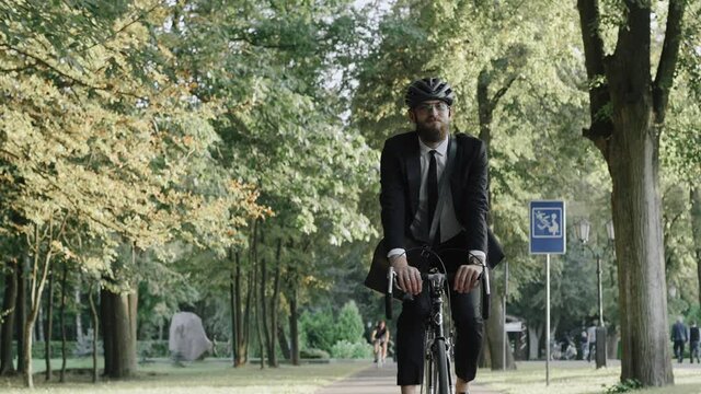Businessman riding a bike in city park wearing suit and helmet, front view. Businessman riding a bicycle in a public park at the sunset. High quality 4k footage