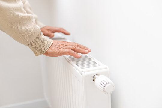 An elderly man warms his hands over an electric heater. In the off-season, central heating is delayed, so people have to buy additional heaters to keep houses warm despite increased electricity bills