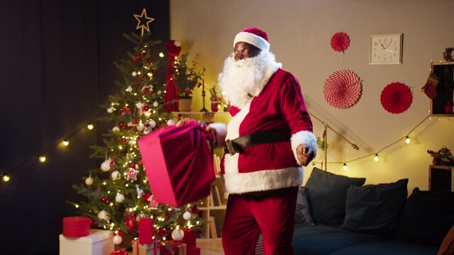 Santa Claus dancing in living room, holding big bag full of gifts. African american man in costume of Santa having fun, showing dance. New year time and christmas holidays concept. 