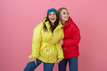 two attractive girl friends active women posing on pink background in colorful winter down jacket...