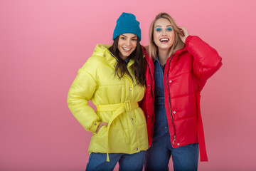 two attractive girl friends active women posing on pink background in colorful winter down jacket...