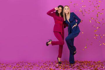two attractive women celebrating new year on violet background in stylish colorful evening suits of purple and blue color, friends having fun together, fashion trend, golden confetti party mood