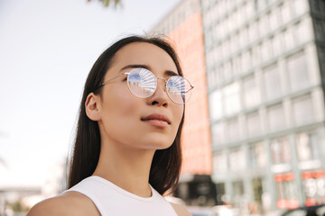 Portrait of fair-skinned young asian woman with confident look up against blurred background. She has dark, straight hair, transparent fashionable glasses, and white sleeveless jacket.