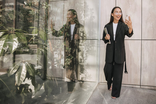 Young asian girl blogger broadcasts live from phone near mirror wall in full growth. She smiles at her followers, dressed in black bell-bottomed trousers, long blazer with belt and open shoes.