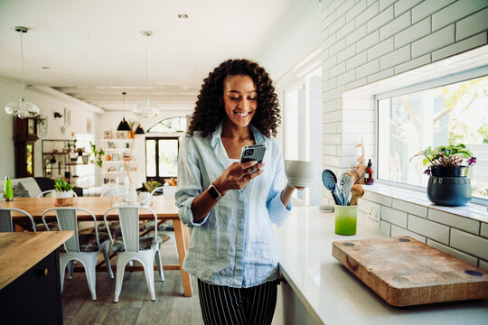 Woman chatting on cellphone standing in kitchen with coffee mug 