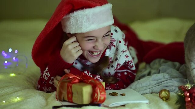 A teenage girl in Santa's red hat and red christmas jacket lies on the bed and writes a letter to Santa Claus. Near the British cat. New Year. hildren's Christmas. Atmosphere. Home.