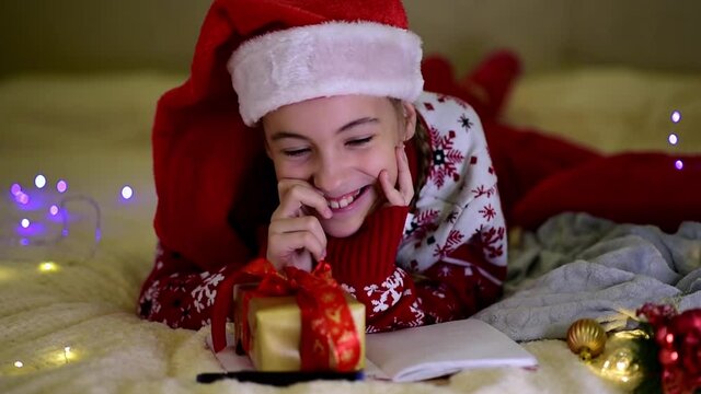 A teenage girl in Santa's red hat and red christmas jacket lies on the bed and writes a letter to Santa Claus. Near the British cat. New Year. hildren's Christmas. Atmosphere. Home.