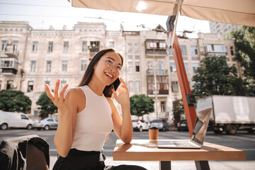 Smiling young asian woman with glasses laughs while talking on phone in street cafe. Laptop and glass of latte are on small table. She sits on chair waving her hand to side.