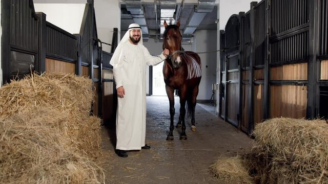 Full length view of the arabian sheikh in kandura stroking his chestnut horse while taking care of him at the stable
