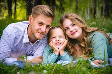 Happy young family father, mother and little daughter smiling as they lie on the green grass on a sunny day