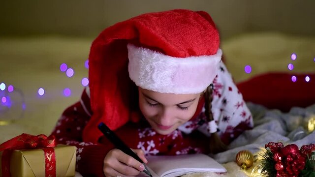 A teenage girl in Santa's red hat and red christmas jacket lies on the bed and writes a letter to Santa Claus. New Year. hildren's Christmas. Atmosphere. Home.
