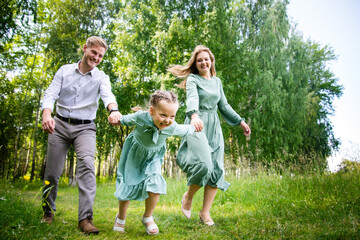 Fototapeta premium Happy young family: father, mother and little daughter smiling, holding hands and walking together on the green grass on a sunny day