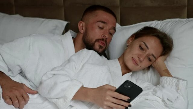 man and woman in bed in morning, lady is viewing social nets in smartphone
