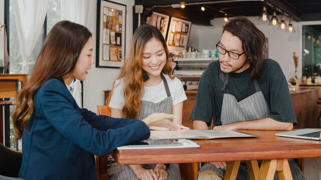 Cheerful Asian Small Business Owners Couple Meeting With Consultant Talking About Business Plan And Finance In Cafe. Young Japanese Agent Consider Signing Contract Insurance Together At Restaurant.