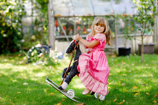 Little preschool girl hugging with rocking horse toy. Happy child in princess dress on sunny summer day in garden. Girl in love with her favourite old vintage toy animal.