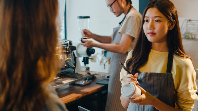 Young Asia female barista waiter taking order from customer standing behind bar counter while talking with customer making note on takeaway coffee cup at cafe restaurant. Owner small business concept.