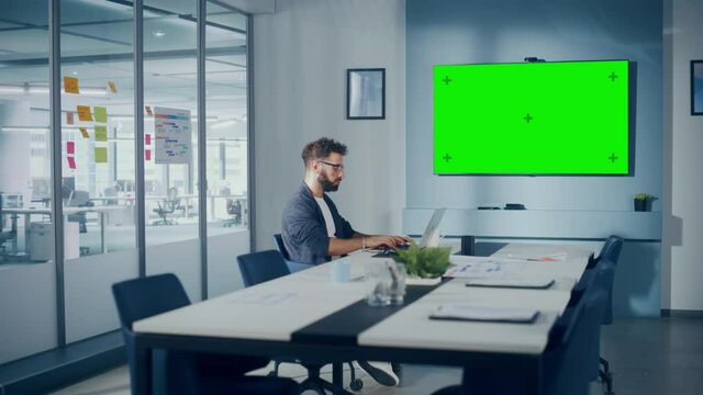 Office Conference Meeting Room: Handsome Hispanic Specialist Working on Laptop Computer interacts with Green Screen Choma Key Wall TV. Businessperson Working in Growing e-Commerce Startup. Wide Shot