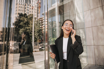 Long-haired young asian brunette has cheerful conversation on phone going to workplace in office. Active lifestyle of business woman. Black formal suit with stripes on her.