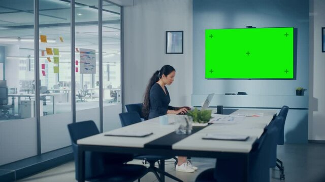 Office Conference Meeting Room: Beautiful Asian Specialist Working on Laptop Computer interacts with Green Screen Choma Key Wall TV. Businessperson Working in Growing e-Commerce Startup. Wide Shot