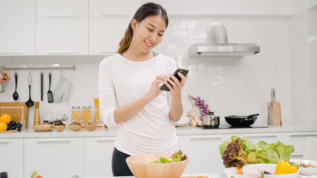 Young Asian blogger woman using smartphone photo post in social media in the kitchen, female making salad at home. Lifestyle women relax at home concept.