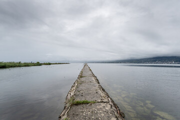 Dike or pier in a lake leading to a point in the distance with a cloudy sky.