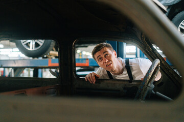 Front view of frustrated young service man in uniform inspecting interior of old car in auto repair shop garage with vehicle background. Concept of car service, repair and maintenance.