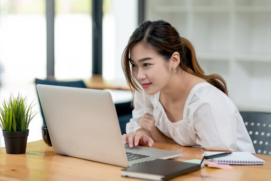 Image of young Asian businesswoman sitting at the office looking at a laptop screen with documents at the desk.