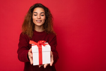 Fototapeta premium Attractive positive smiling young brunette curly woman isolated over red background wall wearing red sweater holding gift box looking at present white box with red ribbon