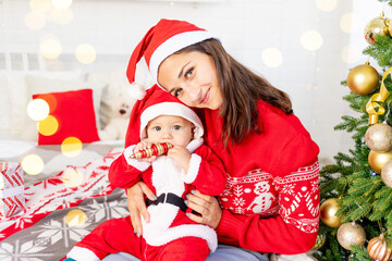 New Year or Christmas, a young mother with a baby on the bed at home by the Christmas tree in a Santa Claus costume hugging and waiting for the holiday smiling