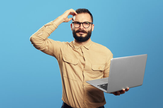 Confused young bearded man holding laptop computer and scratching his head isolated over blue background 