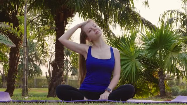 Slow Motion of Fitness Woman Stretching Body, Tilts Head to Side, Stretch Neck on Yoga Mat in City Park. Female Practicing Sport Outdoor, Warm Up Before Workout in Tropical Park. Healthy Lifestyle