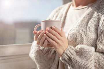 A woman in a warm cardigan by the window holds a mug in her hands. Close-up of female hands holding a cup of coffee or tea.