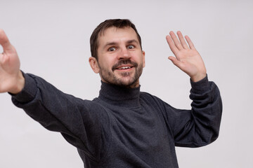 A man with a beard waves at a mobile phone camera during a video chat, talks to someone in an Internet stream, smiles and greets, standing on a white background.