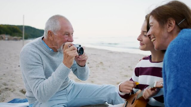 Happy senior couple with granddaughter on beach, having a picnic and taking pictures.