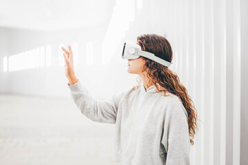 Curly haired young woman in virtual reality glasses moves hands touching online object and looks...
