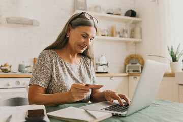 Happy senior woman making online payments of bill using laptop, smiling while shopping with credit card, using application for Internet banking, isolated against kitchen surroundings - obrazy, fototapety, plakaty
