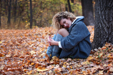 A young girl in gray coat and knitted socks sits in an autumn park. The girl has curly hair. Yellow...