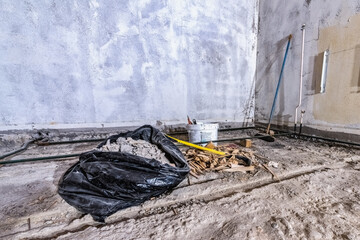 Construction waste debris in black plastic bag and wood chips on old concrete floor, from demolished room, broom, crowbar. Room corner, wide view