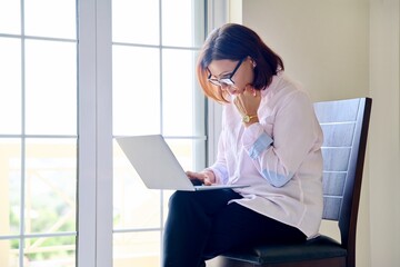 Mature businesswoman in glasses with laptop in her hands near window