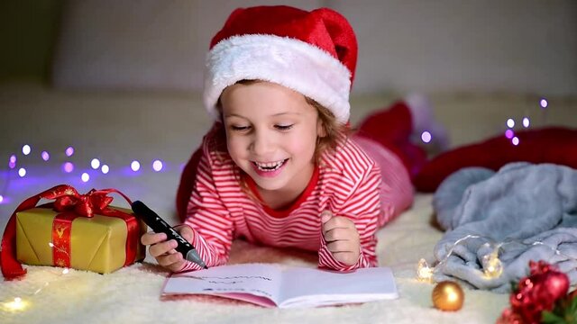 A little smiling girl in Santa's red hat and red christmas clothing lies on the bed and writes a letter to Santa Claus. New Year. Children's Christmas. Atmosphere. Home.