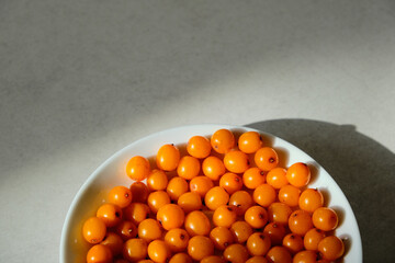 Organic sea buckthorn in a white plate. Layout on a gray table, flat lay. Fresh orange berries in bright sunlight. Natural valuable food supplement.