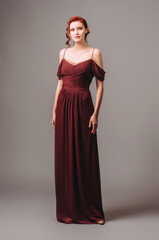 Fototapeta premium Beautiful skinny ginger girl in a burgundy evening gown. Maroon red dress with off the shoulder sleeves. Studio portrait of young lady on grey background.