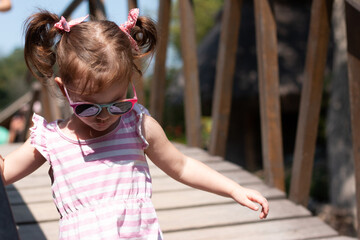 Little beautiful girl with red pigtails and sunglasses walks in a striped dress on a wooden bridge