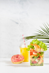 Wellness summer food take away set of orange juice, pink donut, tropical salad with shrimps, lettuce, red pepper, croutons in plastic box in white bar interior with marble tile, palm leaf, vertical.
