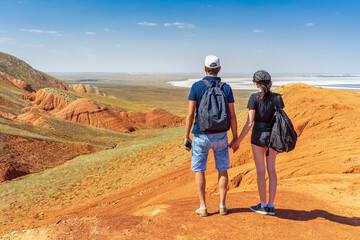 Obraz na płótnie Canvas Family couple travelers walking in mountain area. Rear view of man and woman standing on trail against Martian landscapes of red hills. Bogdo Baskunchak Nature Reserve, Astrakhan region, Russia