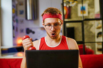 Funny young man geek with dumbbells watching training program on laptop online. Sports and humor...