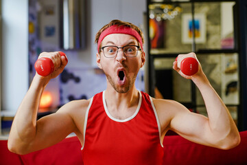Portrait of funny geek exercising and doing sport training. Crazy athletic man in red sportwear using small dumbbells for bodybuilding.