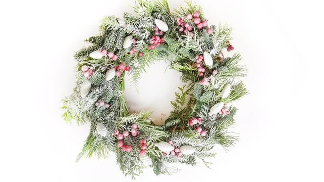 Slow motion looping rotate of snowy Christmas wreath with natural evergreen branches and holly berry on white background. Xmas traditional decorations. 4k video.