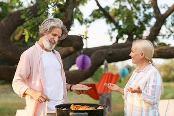 Happy elderly couple cooking food at barbecue party on summer day