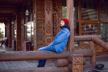 man with a hipster beard in a red knitted hat and a denim jacket is sitting on the stairs at wooden...